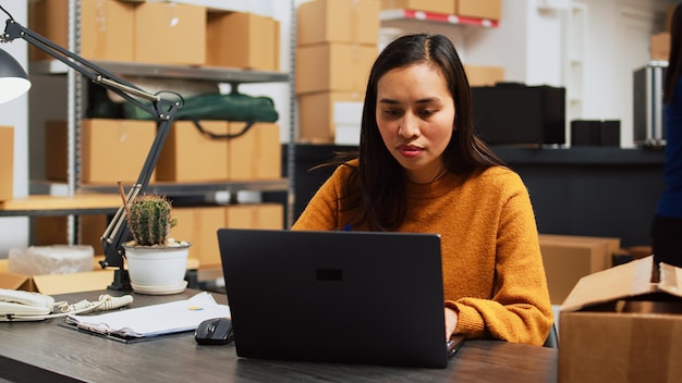 A close-up of a Colombian entrepreneur working on a laptop, surrounded by packages and shipping labels, with a smartphone displaying an e-commerce app. The image highlights the integration of technology and entrepreneurship.